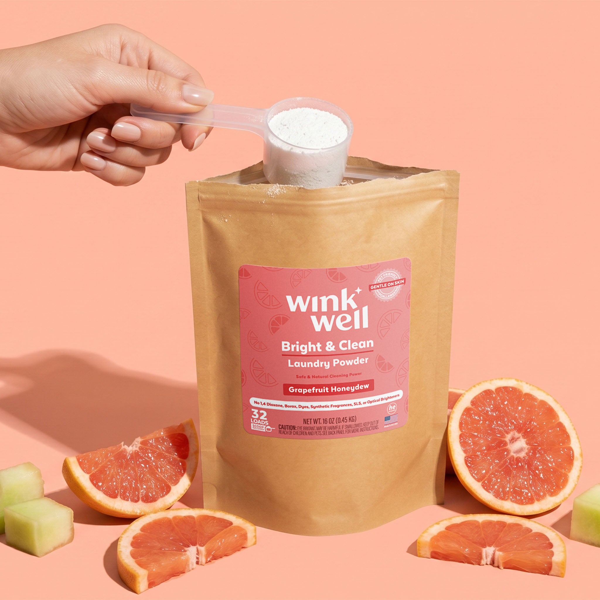 A Wink Well Grapefruit Honeydew Bright & Clean Laundry Powder package with a hand scooping powder, surrounded by grapefruit and honeydew slices on a pink background