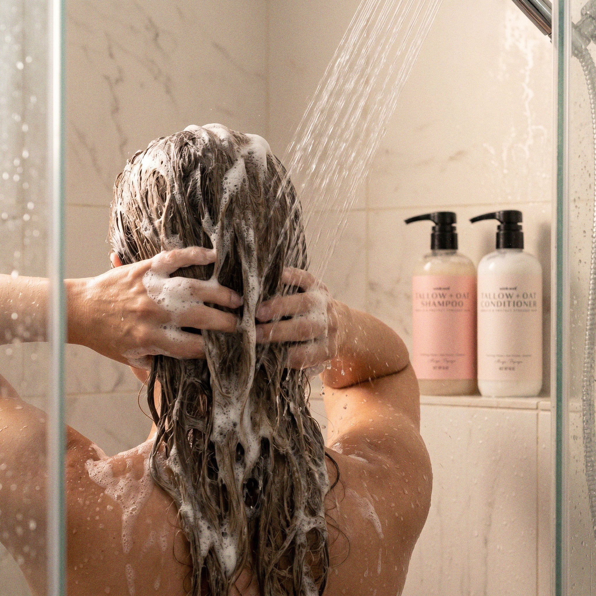 Person washing hair with shampoo in a shower, with bottles of shampoo and conditioner visible.