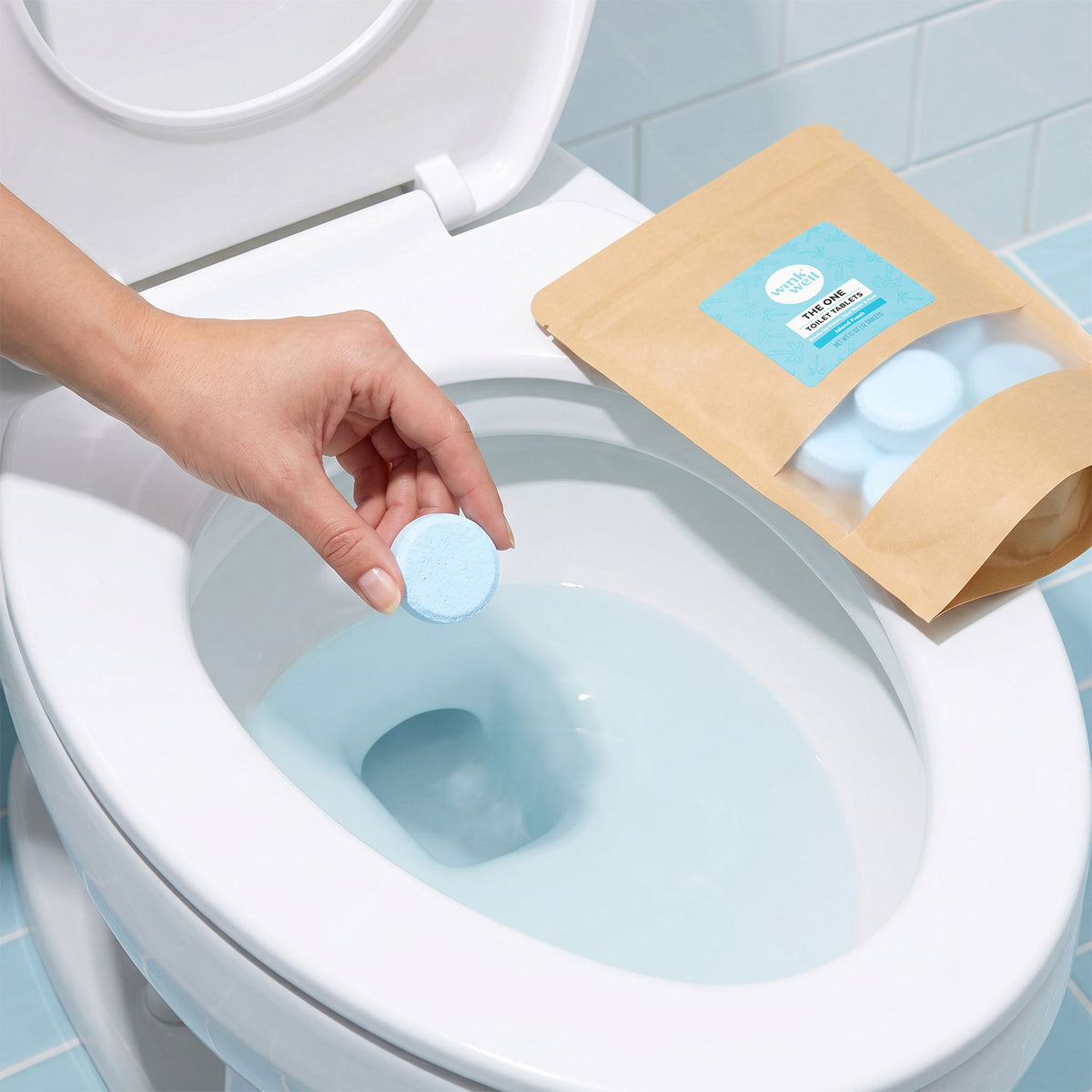 Person placing a blue Wink Well Toilet Tablet into a toilet with a package of Wink Well Toilet Tablets nearby.
