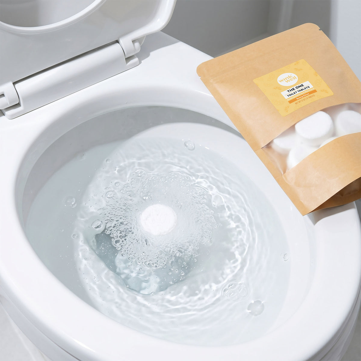 White Wink Well Sunny Citrus toilet tablet fizzing and dissolving in a toilet bowl, with the kraft package resting on the toilet seat