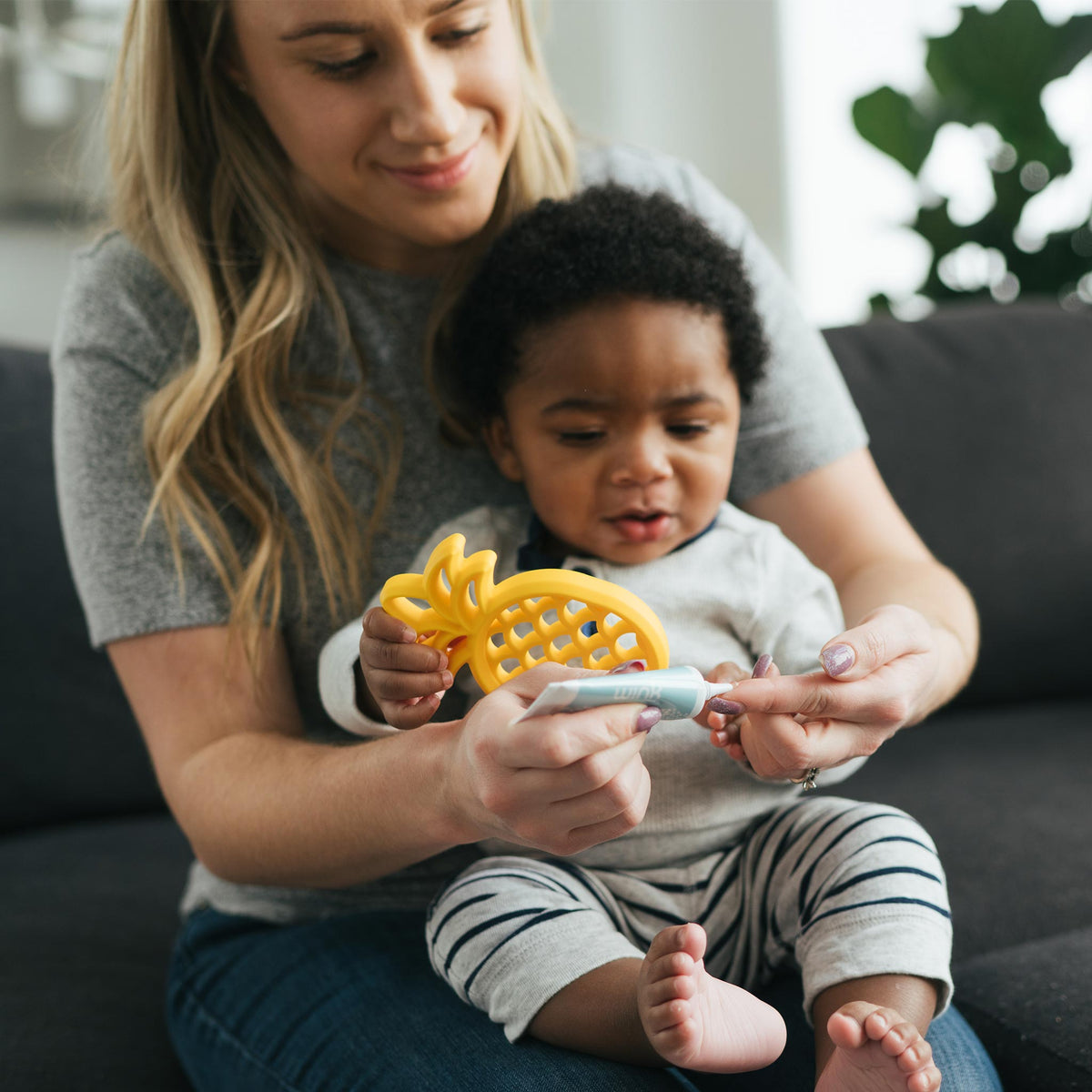 Blonde mother holding a baby with a yellow pineapple teether and Wink Kids Cool Gums Teething Gel
