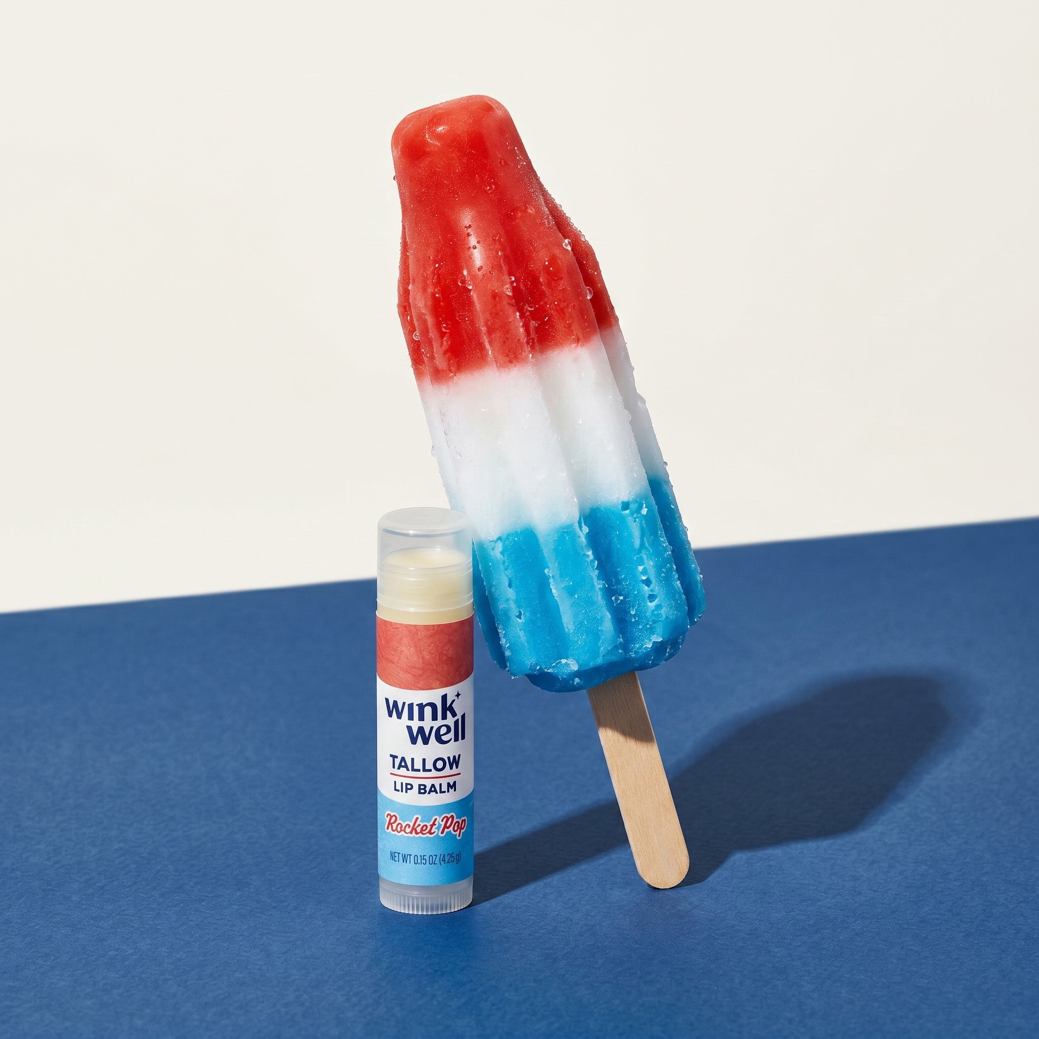 Wink Well Rocket Pop flavored tallow lip balm stick, with a cold bomb pop popsicle leaning against it