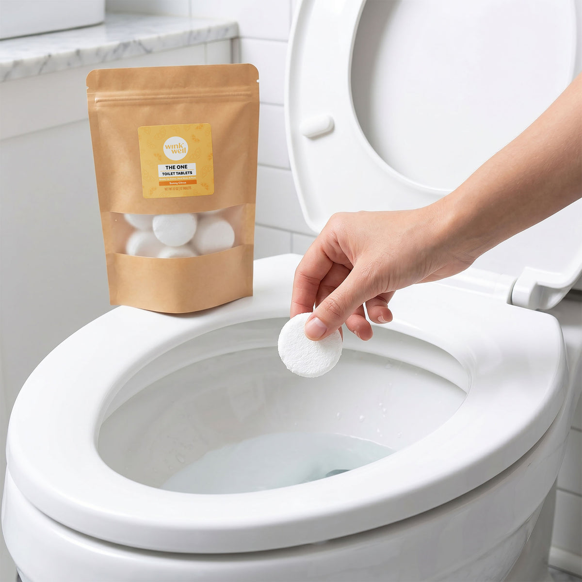 Hand dropping a white Wink Well Sunny Citrus toilet tablet into a toilet bowl, with the kraft package resting on the open seat in a white bathroom