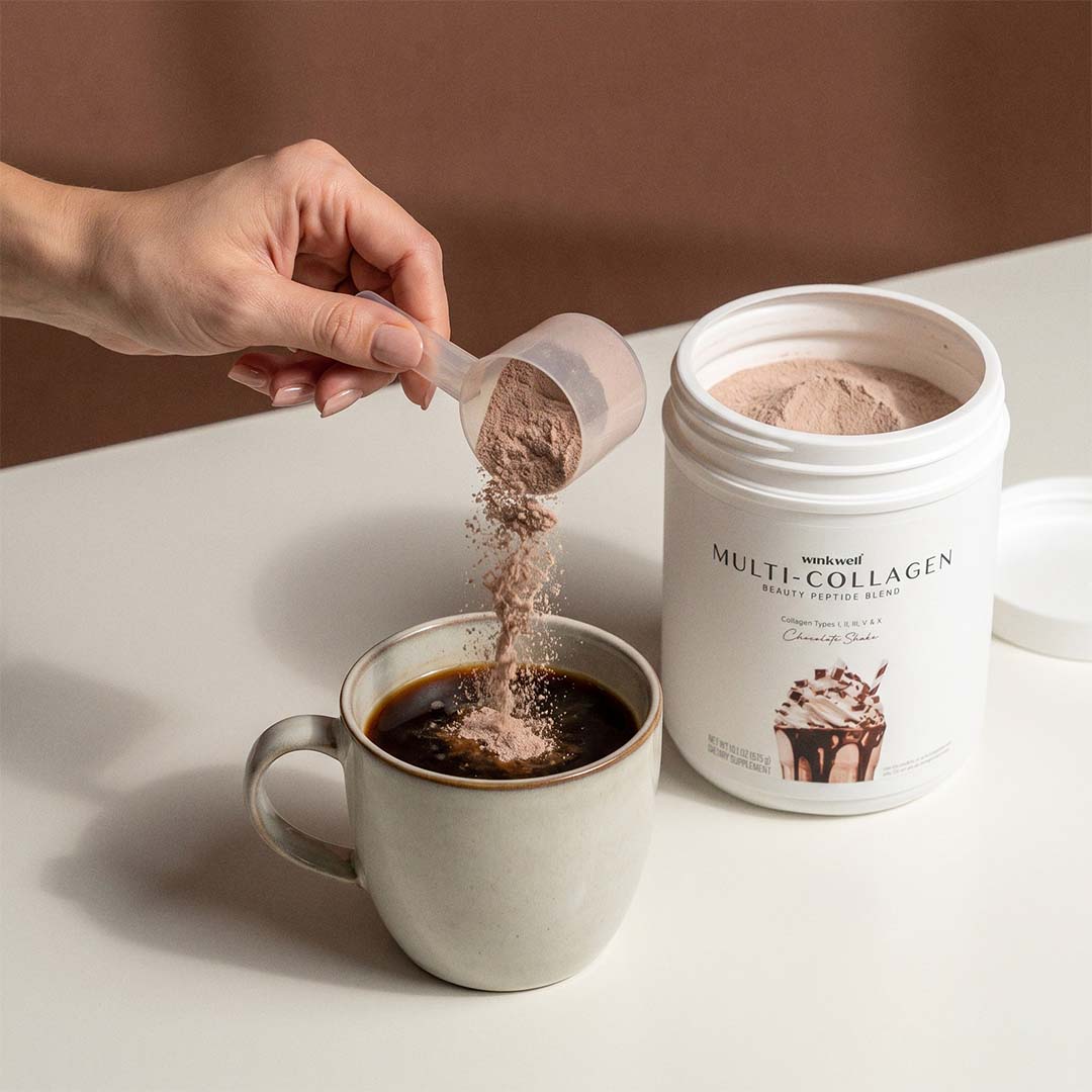 A hand pours Wink Well chocolate collagen from a small scoop into a mug of coffee, with a white tub of Multi-Collagen beside it on a white tabletop against a brown background.